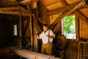 A person in historical attire is giving a demonstration or presentation in a rustic wooden interior, possibly a historic or educational setting.