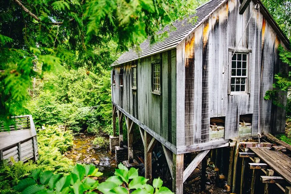 An old wooden building on stilts overlooks a serene stream surrounded by lush greenery