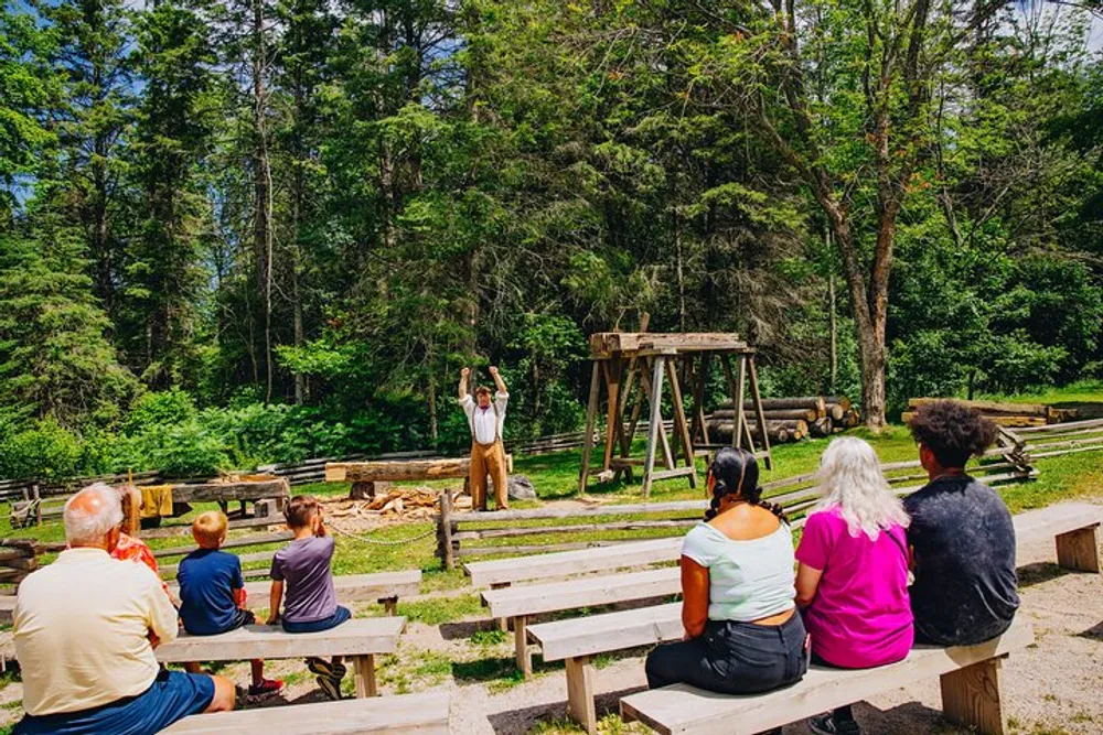 A group of spectators is seated on wooden benches outdoors attentively watching a person demonstrating wood cutting in a forested area