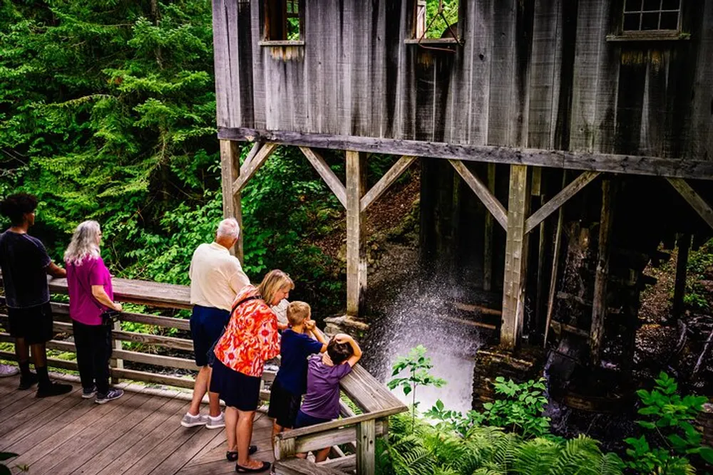 A group of people including children and adults are observing water cascading from an old wooden structure into a stream surrounded by lush greenery