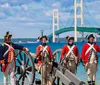 Individuals dressed in historical military uniforms stand beside a cannon with a large suspension bridge in the background