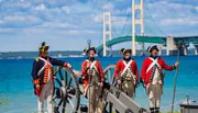 Individuals dressed in historical military uniforms stand beside a cannon with a large suspension bridge in the background.