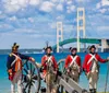 Individuals dressed in historical military uniforms are standing next to a cannon with a modern suspension bridge in the background