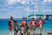 Individuals dressed in historical military uniforms are standing next to a cannon with a modern suspension bridge in the background.