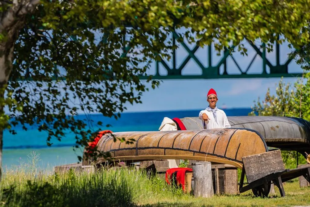 A person wearing a red hat and white outfit is sitting on a collection of overturned canoes near a body of water with greenery and a bridge structure in the background