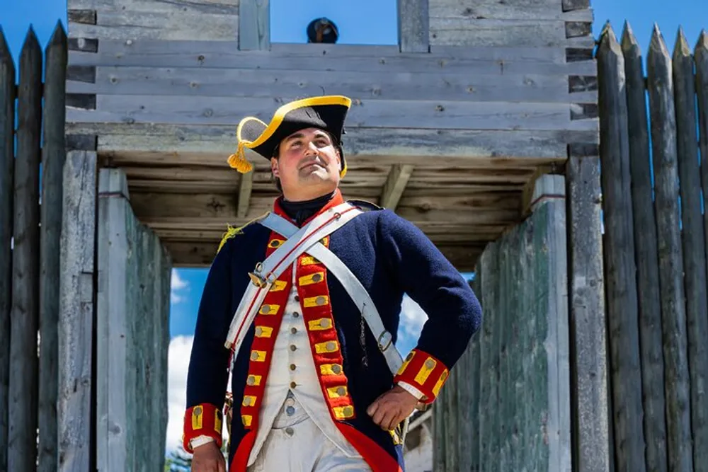 A person dressed in a historical military uniform stands confidently with their hands on their hips against a backdrop of a wooden palisade and watchtower under a clear blue sky