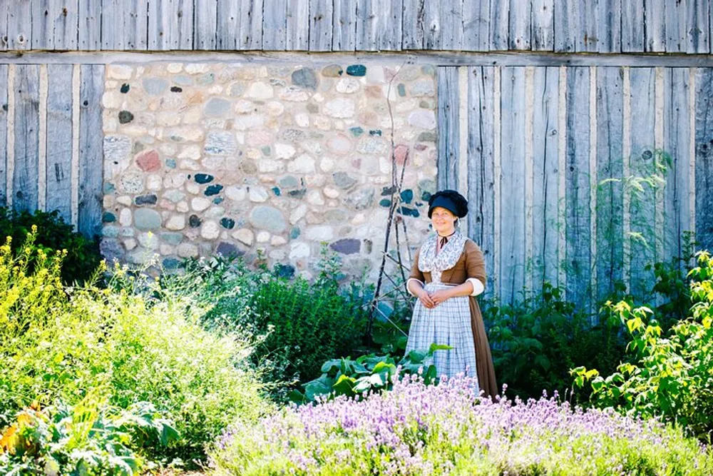 A person in historical costume stands in a sunny garden in front of a rustic stone and wood barn