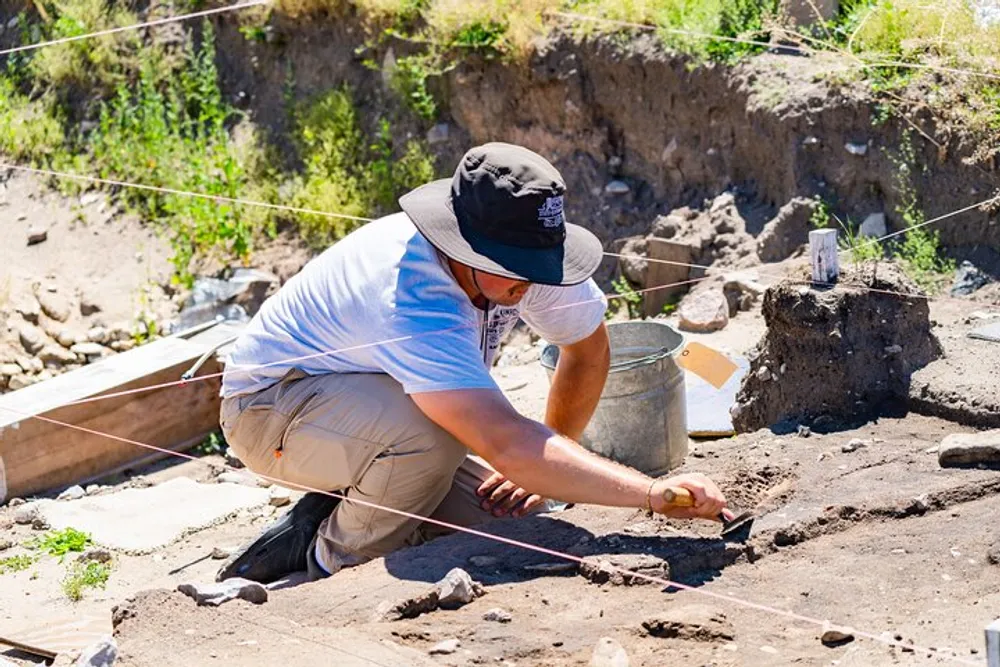 An archaeologist meticulously excavates a site using a brush near a grid of strings to uncover artifacts in the earth