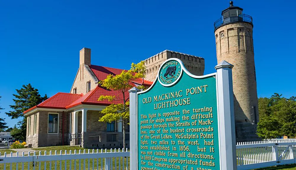 The image shows the historical Old Mackinac Point Lighthouse and its associated buildings under a clear blue sky accompanied by an informational sign