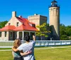 A couple is enjoying a sunny day outdoors while the man points at a historic lighthouse and adjacent building possibly discussing the site or preparing to take a photo