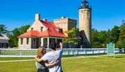 A couple is enjoying a sunny day outdoors while the man points at a historic lighthouse and adjacent building, possibly discussing the site or preparing to take a photo.