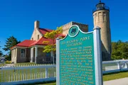 The image features the Old Mackinac Point Lighthouse and its informative sign under a clear blue sky.