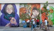 A group of people is seen in front of a vibrant street mural featuring an ethereal figure with a galaxy within her hands, juxtaposed with other colorful and fantastical elements.