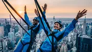 Two individuals in safety harnesses are gleefully experiencing an urban high-altitude adventure against a backdrop of a city skyline at sunset.