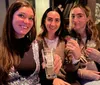 Three women are smiling and holding drinks appearing to enjoy a social gathering indoors