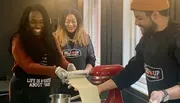 Three people are enjoying a cooking class, using a pasta maker to roll out dough, with aprons on that read 