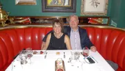 A couple is sitting at a well-set table with red upholstery in a restaurant, smiling at the camera with wine glasses and dessert in front of them.