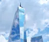 The image shows a tall skyscraper with a reflective glass facade reaching into a cloudy blue sky surrounded by other buildings