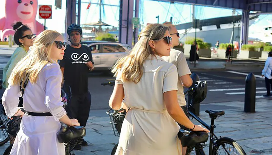 The image captures a moment on a sunny street where two women appear to be getting ready for a bike ride, with other people and a whimsical pink figure in the background.