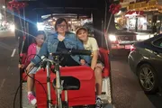 Three people, likely part of a family, are sitting in a pedicab at night on a city street, smiling for the photograph.