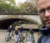 A man takes a selfie in the foreground with four people with bicycles standing under a bridge in the background