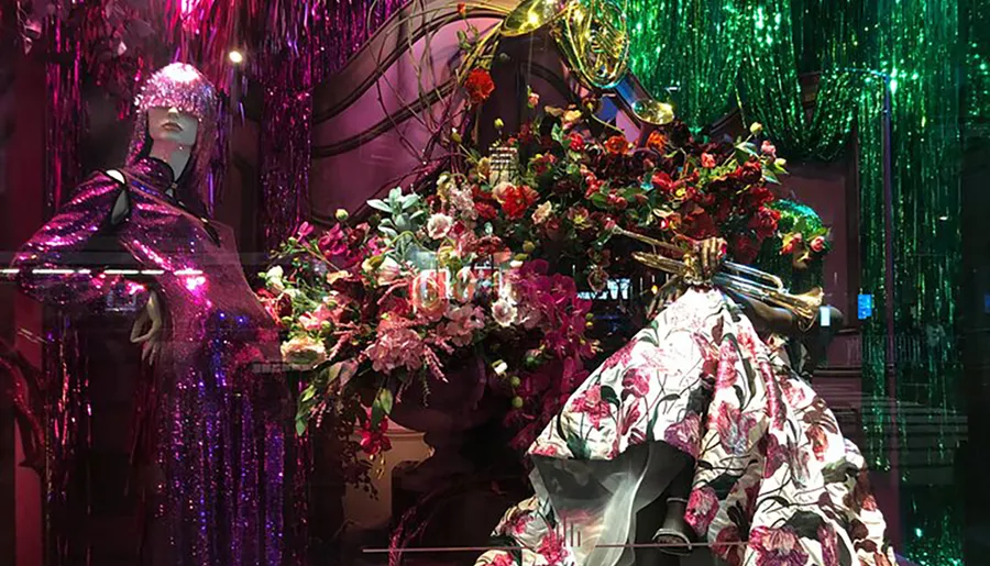 A vivid and intricate window display featuring a mannequin dressed in a floral gown and a sequined headpiece, surrounded by an abundance of flowers and shimmering decor.