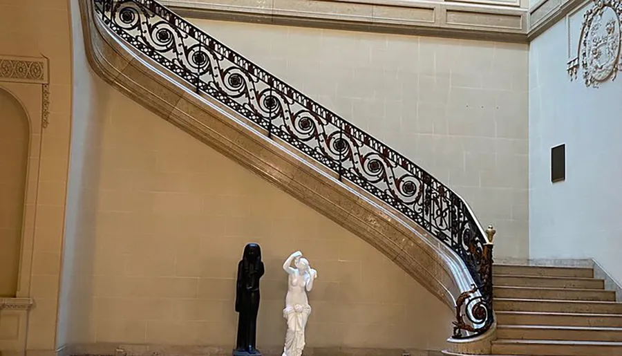 An elegant staircase with intricate ironwork on the banister is flanked by two sculptures, creating a sophisticated and artistic setting.