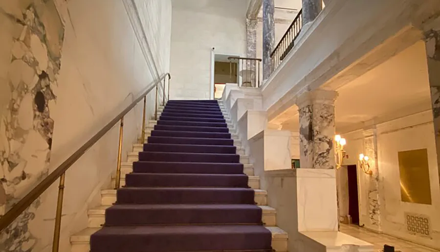 This image shows an elegant staircase with purple carpeting, marble accents, and wall sconces, exuding a blend of luxury and historical charm.