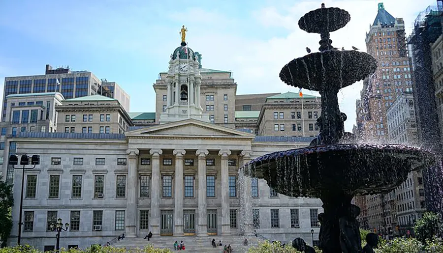 An ornate fountain is in the foreground with the Brooklyn Borough Hall and surrounding buildings in the background on a sunny day.