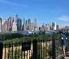 Two people are admiring the view of a city skyline from a park with a No Bicycling sign on the fence