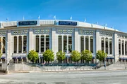 The image shows the exterior of Yankee Stadium, a prominent baseball park, on a sunny day with clear blue skies.