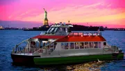 A ferry cruises near the Statue of Liberty during a vibrant sunset.
