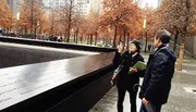 Two individuals are standing by a memorial with inscribed names, pointing and appearing to have a discussion.