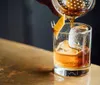 A cocktail is being poured into a glass containing a large ice sphere garnished with a twist of citrus peel secured by a mini clothespin