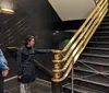 Two people are standing near a staircase with black steps and a golden railing inside a building