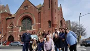 A group of people is posing for a photo on a sunny day in front of a red-brick church with a large rose window.