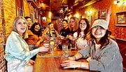 A group of people is happily socializing around a long table inside a cozy bar or restaurant with drinks in front of them.