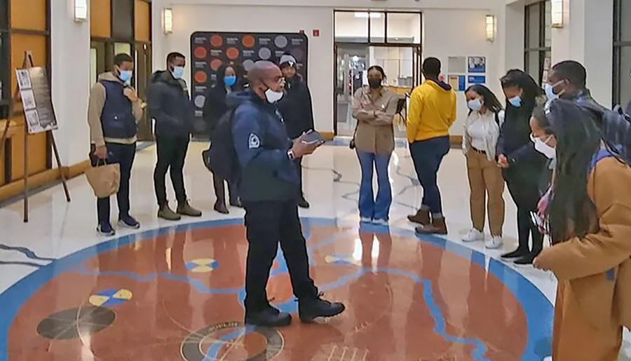 A group of people, some wearing masks, are listening to a person speaking in the center of a lobby with a decorative floor design.