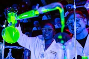 Two people wearing lab coats and safety glasses are working with glowing green chemicals in a laboratory setting.