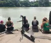 Four individuals are sitting by a lake in a park with one pointing towards the water while an electric scooter rests on the ground beside them