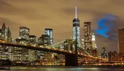 The image shows a nighttime view of the Brooklyn Bridge with the illuminated Manhattan skyline in the background.