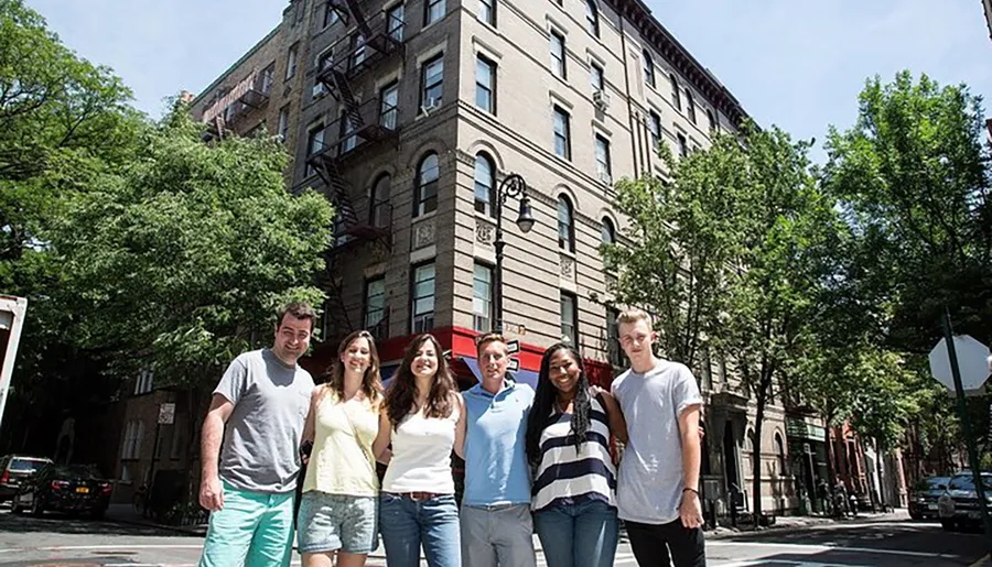 A group of six individuals is posing for a photo on a sunny day in front of an old brick building with fire escapes and trees lining the street.