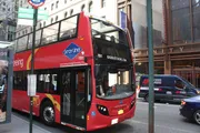 A red double-decker sightseeing tour bus is parked on a city street with cars passing by and urban buildings in the background.