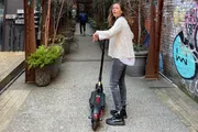 A person stands on an electric scooter in a narrow urban alleyway, turning to look at the camera with a mild expression.