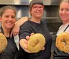 Three smiling people are holding up large seeded bagels toward the camera in a kitchen setting