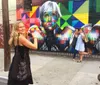 A woman is posing in front of a vibrant street mural with another woman playfully photo-bombing in the background