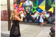 A woman is posing in front of a vibrant street mural with another woman playfully photo-bombing in the background.