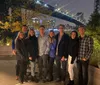 A group of smiling people pose for a photo at night with a lit-up bridge and city skyline in the background