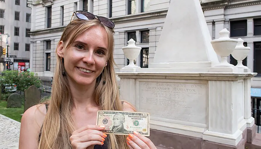 A smiling woman is holding a ten-dollar bill in front of Alexander Hamilton's grave.