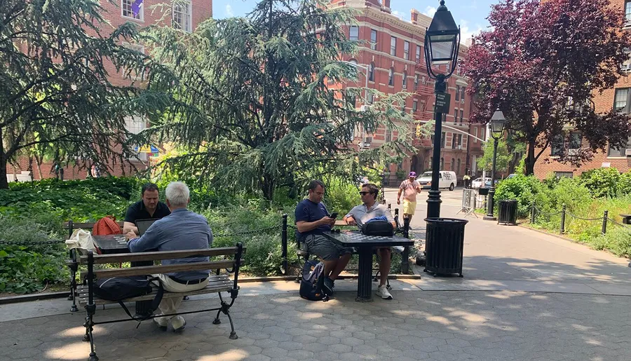 People are enjoying a sunny day in a park with benches, surrounded by lush greenery and a backdrop of classic brick buildings.